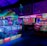 An indoor arcade filled with vibrant gaming machines lit with neon blue and pink lights. The scene includes various arcade games with flashing screens and colorful displays, and a large overhead structure resembling a green snake. The background shows illuminated signs and themed decor, contributing to an immersive entertainment atmosphere.