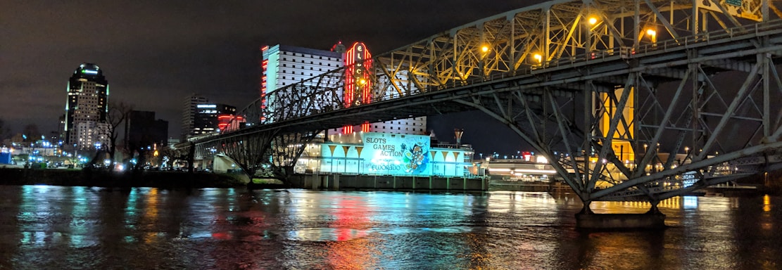 A brightly lit casino with neon signs and red lights is reflected in a river. A bridge stretches across the water, with an urban skyline visible in the background against an overcast night sky.