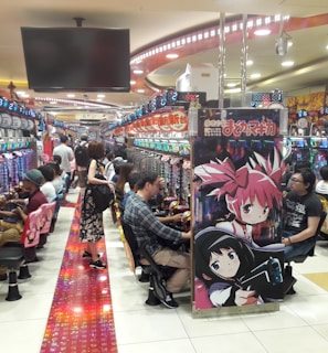 A bustling arcade filled with people playing various gaming machines. Brightly colored displays and animated characters are prominent on posters and screens. The atmosphere is lively with patrons seated in rows, focusing on the games. Overhead signage and electronic scoreboards are visible, contributing to the busy and vibrant environment.