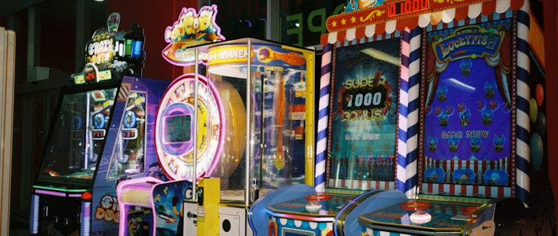 A row of brightly colored arcade game machines is arranged in a line against the wall. Each machine features vibrant lights and designs, with themes ranging from claw games to game show-inspired setups. The area has a playful and inviting atmosphere, suitable for entertainment or amusement centers.